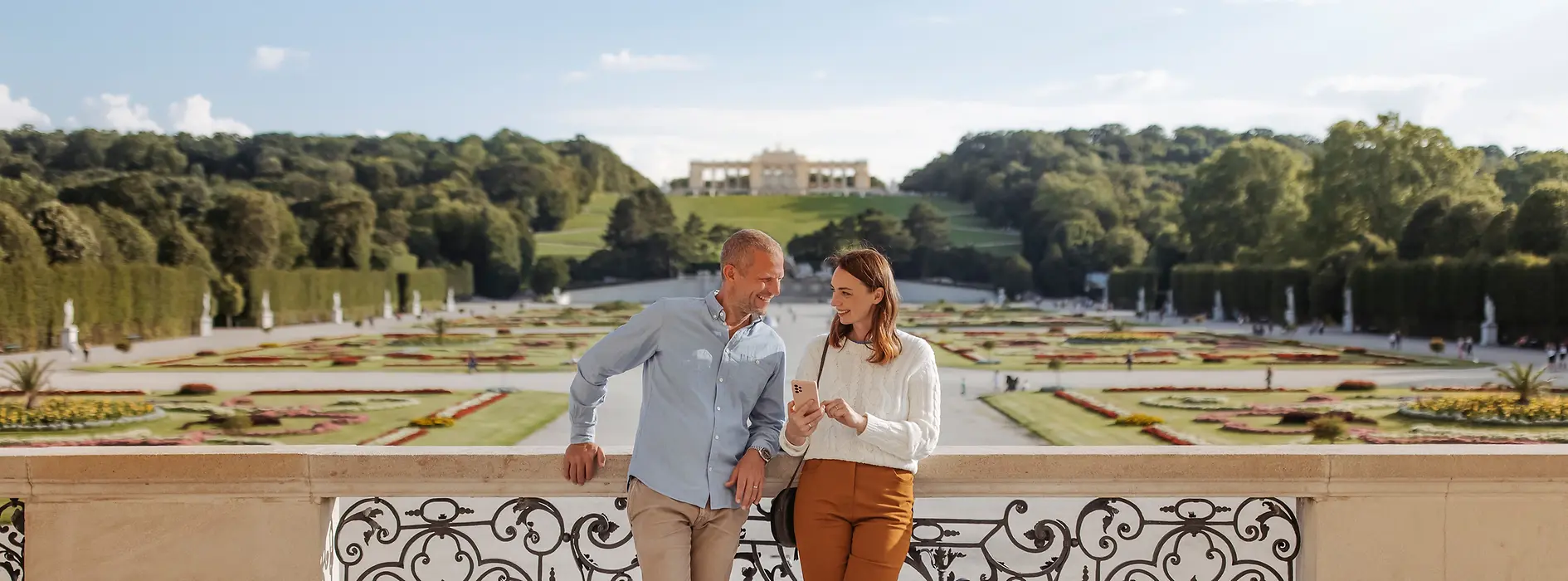 Ein Paar schaut gemeinsam aufs Handy im Hintergrund der Schloss Schönbrunn Garten und die Gloriette