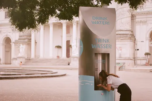Frau trinkt an einem Trinkbrunnen, im Hintergrund die Karlskirche